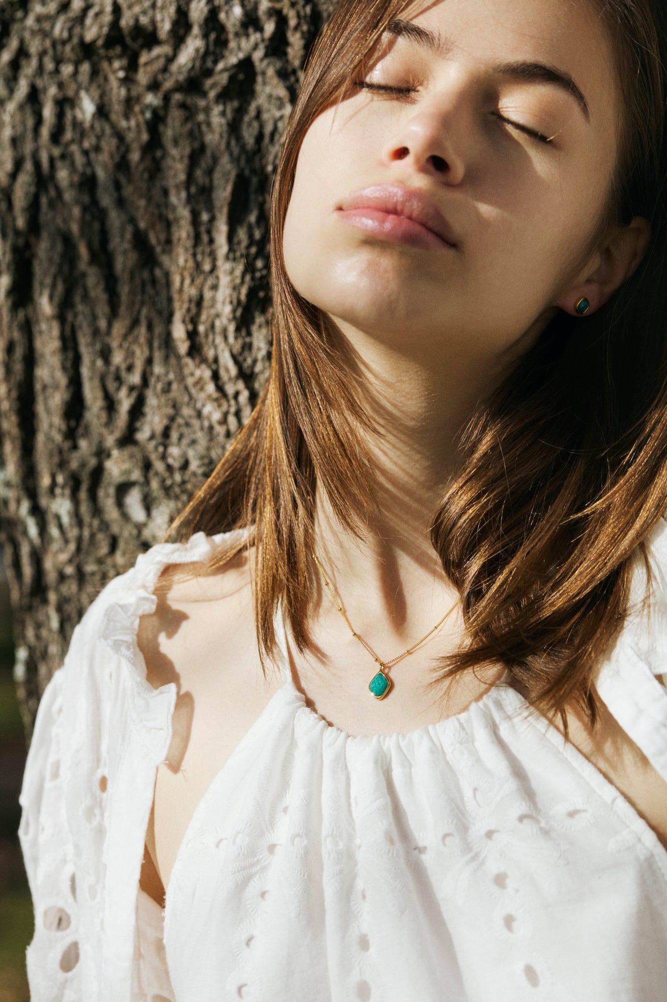 Woman with closed eyes leaning against a tree, wearing a white blouse and a necklace with an amazonite pendant.